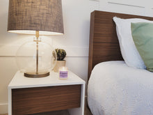 Load image into Gallery viewer, The Milkshake candle is on a bedside table in a fancy decorated bedroom. There is a nice lamp, a succulent, and a nicely made bed.