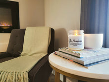 Load image into Gallery viewer, There is a candle called The Pandemic sitting on books on a side table. Beside it is a chair with a blanket and a cozy fireplace.