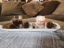 Load image into Gallery viewer, The Pumpkin Candle is sitting on a serving tray on a footrest. The serving tray has a cup of coffee or tea, and decorative fall pinecones. You can see a comfy looking couch in the background.
