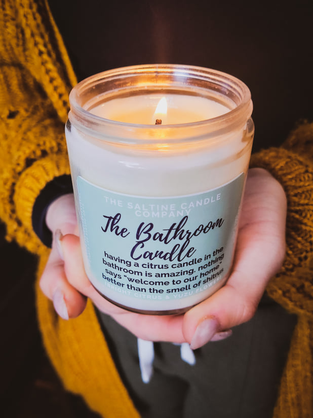 Picture is of a woman with long pretty nails holding a soy wax candle called The Bathroom Candle. The candle is lit and burning.