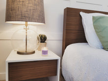 Load image into Gallery viewer, The Milkshake candle is on a bedside table in a fancy decorated bedroom. There is a nice lamp, a succulent, and a nicely made bed.
